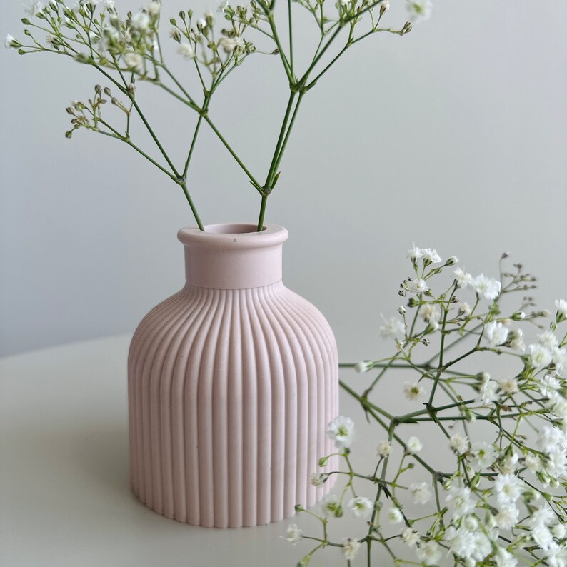 May include: A pale pink ceramic vase with vertical ridges, holding a bouquet of white baby's breath flowers.