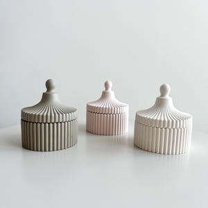 May include: Three decorative storage containers with lids. The containers are made of concrete and have a ribbed design. The containers are in three colours: grey, pink, and white.