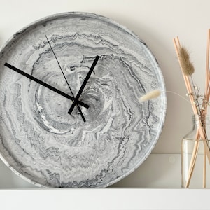May include: A round, grey and white marble clock with black hands. The clock has a swirling pattern and is hanging on a white wall.