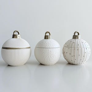 May include: Three white ceramic bauble-shaped containers with silver-coloured metal tops. The containers have different textures: one with vertical lines, one with a textured surface, and one with vertical lines and small silver dots. The containers are on a white surface.