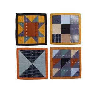以下が含まれることがあります： 青、茶色、黄色の幾何学模様が施された布製のコースター4枚。コースターは黒またはオレンジの縁取りがされています。