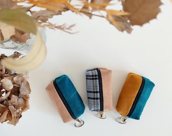 May include: Three small zippered pouches with keyrings. The pouches are made of fabric and have a two-tone design. One pouch is teal and beige, one is plaid and beige, and one is mustard yellow and beige.