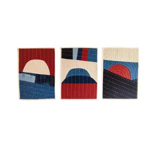 Op de afbeelding: Drie abstracte, gewatteerde wandkleden. Elk heeft een uniek ontwerp met een wit, rood en blauw kleurenpalet. De ontwerpen omvatten geometrische vormen die op landschappen lijken, met gestikte details en een crèmekleurige rand.