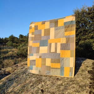 May include: A patchwork quilt with squares of yellow, blue and brown fabric. The quilt is hanging on a large rock in a field.