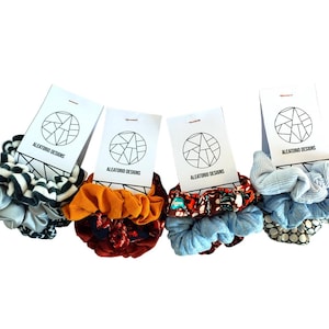 May include: Four sets of hair scrunchies in different patterns and colors. Each set includes two scrunchies. The scrunchies are packaged on white cards with the text "ALEATORIO DESIGNS".