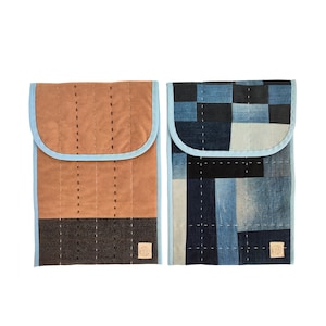 Puede incluir: Dos fundas acolchadas para portátiles. Una es marrón con una base marrón oscuro y ribete azul claro. La otra es de patchwork de mezclilla con ribete azul claro. Ambas tienen un pequeño parche cuadrado con un logotipo.