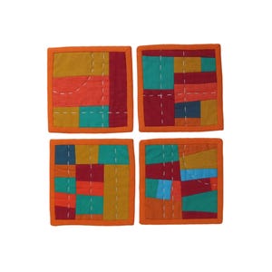 Op de afbeelding: Set van vier kleurrijke stoffen onderzetters met geometrische patchwork-ontwerpen. Elke onderzetter heeft een andere opstelling van rode, oranje, gele, groene en blauwe stofvierkanten.
