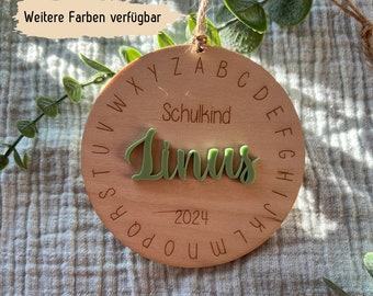 May include: A wooden circle with the alphabet and the year 2024 engraved. The word "Schulkind" and the name "Linus" are written in green on the circle.