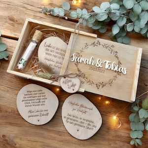 Könnte beinhalten: Eine hölzerne Geschenkbox mit Schiebedeckel, die ein kleines Glasfläschchen mit der Zahl 50 und mehrere herzförmige und ovale Holzplaketten mit Text enthält. Der Deckel ist mit einem floralen Kranz und den Namen "Sarah & Tobias" sowie dem Datum 25.03.2022 graviert.