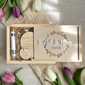 Könnte beinhalten: Eine Holzbox mit Schiebedeckel, mit den Namen "Sarah & Florian" und dem Datum "12.06.2026" beschriftet. Im Inneren befinden sich ein kleines Glasröhrchen mit einem 50-Euro-Schein, ein Holzherz mit "Alles Liebe" und eine runde Holzplakette mit Text.