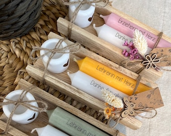 May include: A collection of decorative candles in various colors, including pink, yellow, and green, arranged on wooden trays. Each candle has text printed on it. The trays are decorated with dried flowers and small wooden accents.