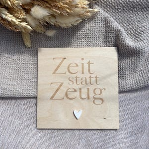 Può includere: Un cartello quadrato in legno con le parole "Zeit statt Zeug" in un carattere marrone chiaro e un piccolo cuore bianco. Il cartello è su una superficie grigia chiara strutturata, con fiori secchi sullo sfondo.