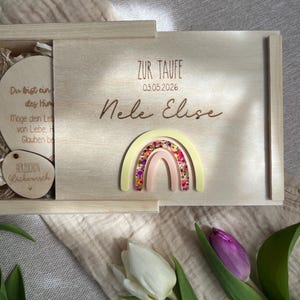 以下が含まれることがあります： スライド式の蓋が付いた明るい木製の箱で、虹と「Nele Elise」の名前で装飾されています。箱には「ZUR TAUFE」の文字と日付03.05.2026が記載されています。箱の中にはテキストが書かれた木のディスクが見えます。