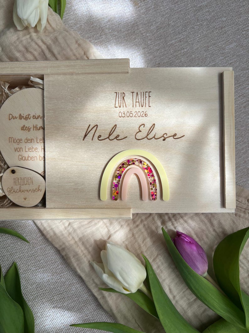以下が含まれることがあります： スライド式の蓋が付いた明るい木製の箱で、虹と「Nele Elise」の名前で装飾されています。箱には「ZUR TAUFE」の文字と日付03.05.2026が記載されています。箱の中にはテキストが書かれた木のディスクが見えます。