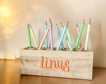 May include: A wooden pencil holder with a light wood finish and white polka dots. The holder has 16 slots for pencils and the name "Linus" is written on the front in orange letters.