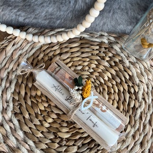 May include: A pair of white candles with the text "Alles Liebe zur Taufe" in a wooden holder, tied with twine. The candles are decorated with a small floral arrangement and a fish symbol. The background includes a woven mat and a gray, furry surface.
