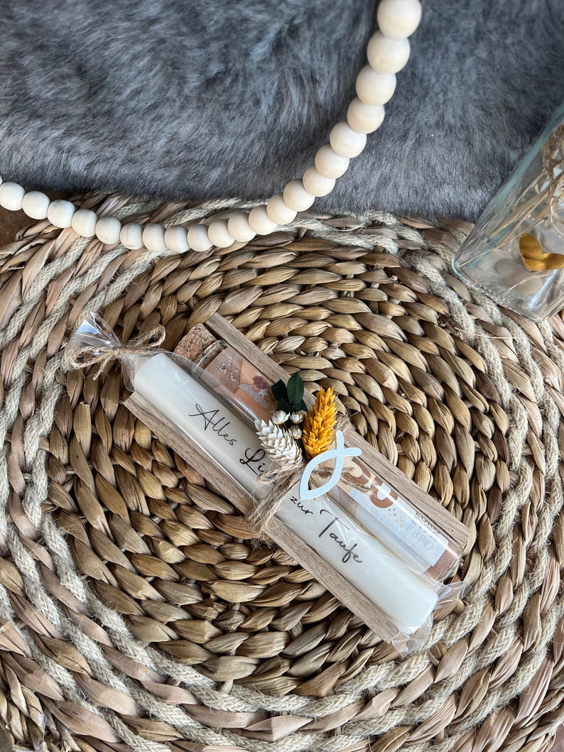 May include: A pair of white candles with the text "Alles Liebe zur Taufe" in a wooden holder, tied with twine. The candles are decorated with a small floral arrangement and a fish symbol. The background includes a woven mat and a gray, furry surface.