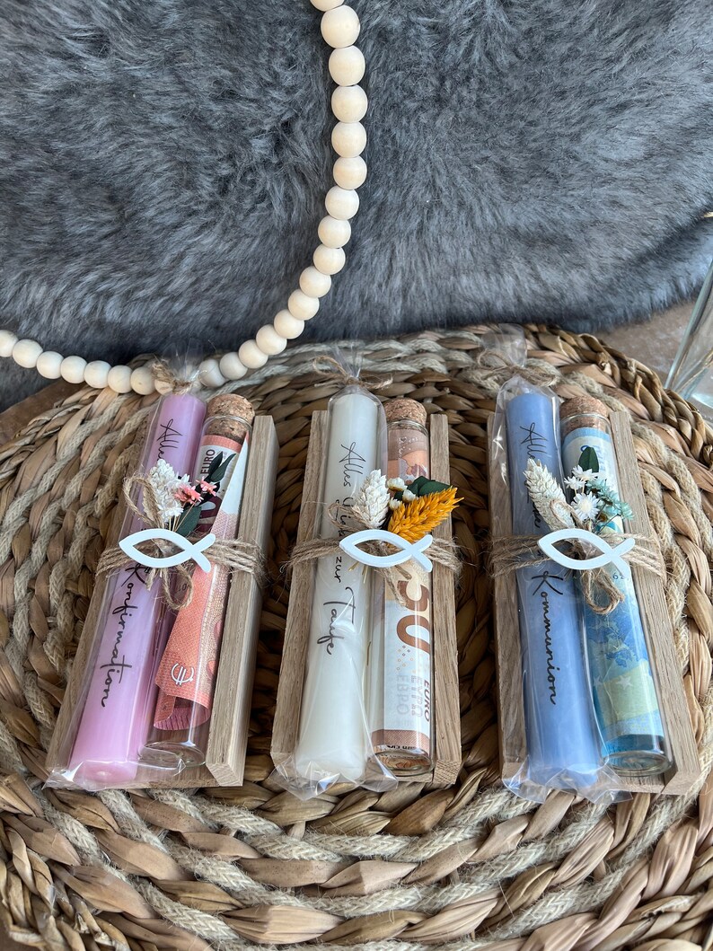May include: Three gift sets in wooden holders, each containing a candle, a small glass bottle, and decorative elements. The candles are in shades of pink, white, and blue. The bottles have cork stoppers and are tied with twine.