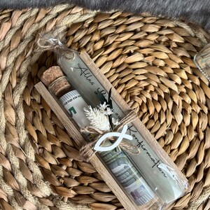 May include: A gift set in a wooden frame, featuring a glass tube with rolled-up currency, a small corked bottle, and a candle with text. The set is decorated with a white fish symbol and dried flowers, all tied with twine.