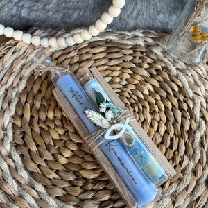 May include: A wooden gift box with two blue candles and a glass vial, decorated with dried flowers and a fish-shaped ornament. The candles are inscribed with the words "Alles" and "Kommunion". The items are arranged on a woven mat.