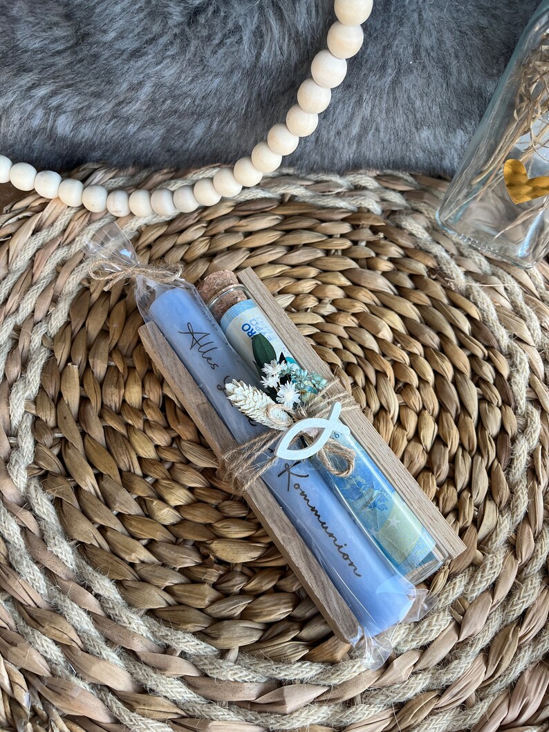 May include: A wooden gift box with two blue candles and a glass vial, decorated with dried flowers and a fish-shaped ornament. The candles are inscribed with the words "Alles" and "Kommunion". The items are arranged on a woven mat.