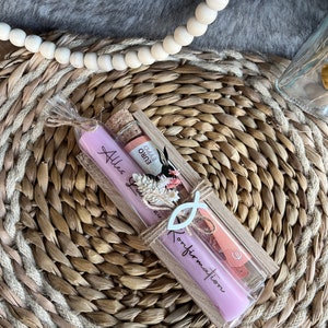 May include: A gift set featuring a lavender candle with the text "Alles Gute zur Konfirmation", a small bottle with a cork, and a decorative fish symbol. The items are packaged in a clear box tied with twine.