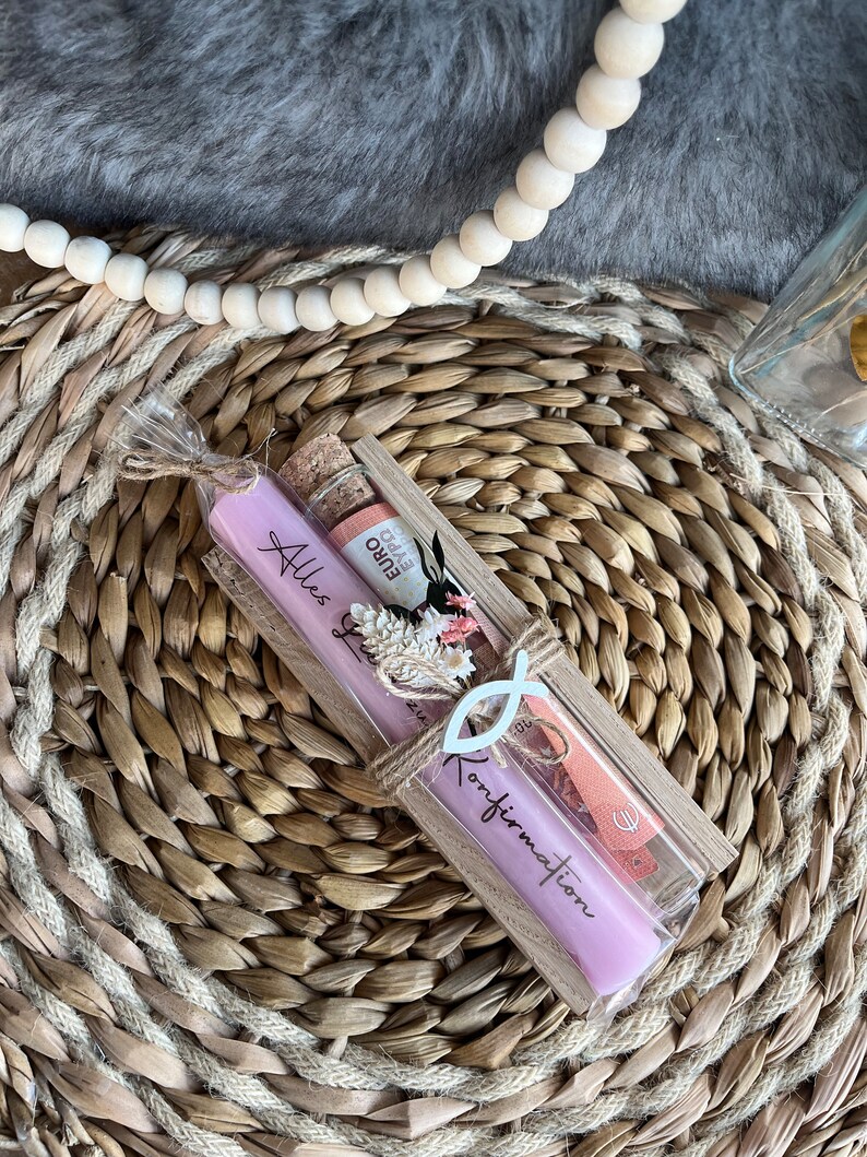 May include: A gift set featuring a lavender candle with the text "Alles Gute zur Konfirmation", a small bottle with a cork, and a decorative fish symbol. The items are packaged in a clear box tied with twine.