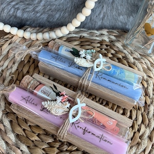 May include: Two long, cylindrical candles in clear packaging, one lavender and one light blue, each with text. The candles are in wooden holders, decorated with twine, dried flowers, and a white fish-shaped charm. Currency is visible in the packaging.