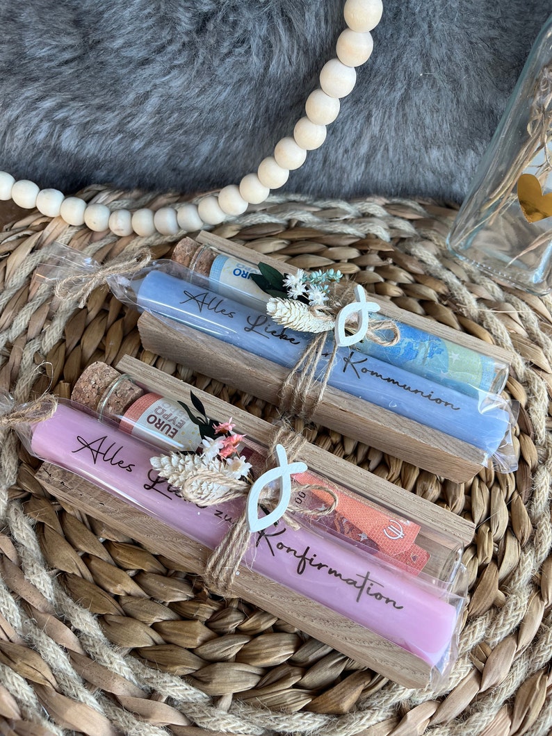 May include: Two long, cylindrical candles in clear packaging, one lavender and one light blue, each with text. The candles are in wooden holders, decorated with twine, dried flowers, and a white fish-shaped charm. Currency is visible in the packaging.