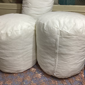 Puede incluir: Tres almohadas cilíndricas blancas, dos de las cuales están sobre una superficie estampada. Las almohadas están hechas de una tela blanca suave y tienen una cremallera en el lateral.
