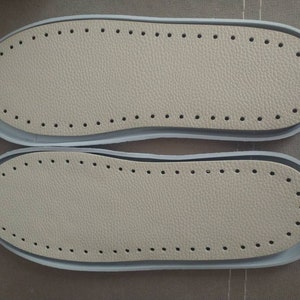May include: Two beige leather shoe soles with a row of holes around the edge. The soles are on a brown surface.