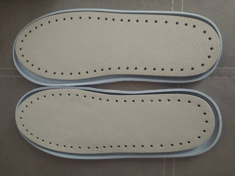 May include: Two beige leather shoe soles with a row of holes around the edge. The soles are on a brown surface.