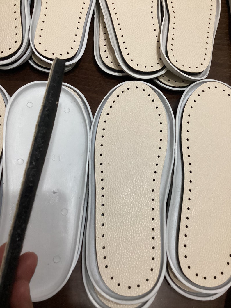 May include: Close-up of several white leather shoe insoles with a black dotted border. The insoles are stacked on top of each other.