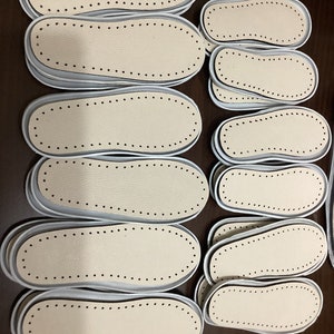 May include: A close-up of several beige leather shoe insoles with white edges and rows of small black dots around the edges. The insoles are stacked on top of each other on a brown surface.