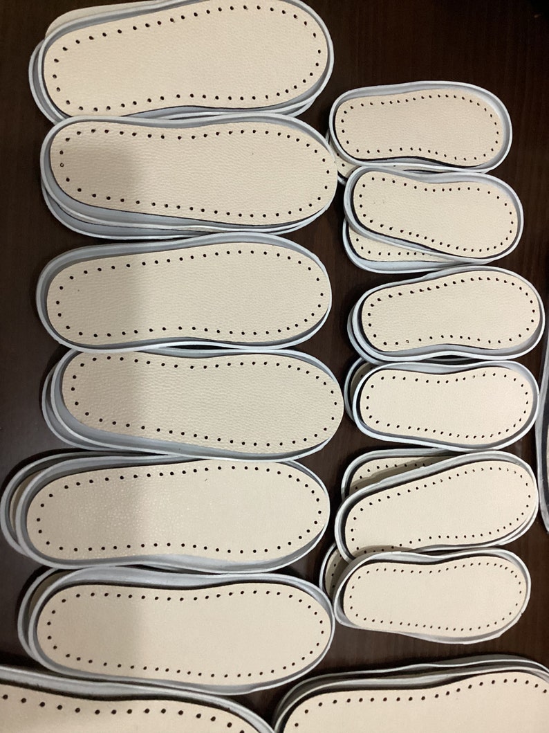 May include: A close-up of several beige leather shoe insoles with white edges and rows of small black dots around the edges. The insoles are stacked on top of each other on a brown surface.