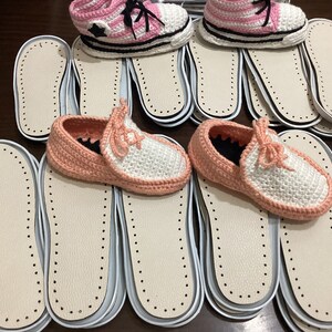 May include: A stack of white and pink crocheted slippers with white soles. The slippers are on top of a stack of white insoles with a dotted pattern.