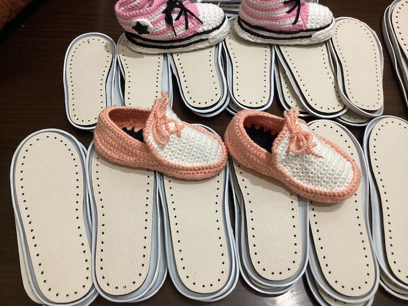 May include: A stack of white and pink crocheted slippers with white soles. The slippers are on top of a stack of white insoles with a dotted pattern.