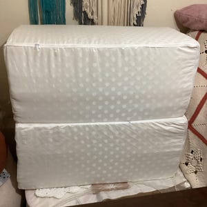 May include: Two white, rectangular cushions stacked. The cushions have a white polka dot pattern and a zip closure. The cushions are on a white surface.