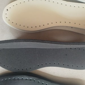 K&ouml;nnte beinhalten: Nahaufnahme von mehreren Schuh-Innensohlen in verschiedenen Farben. Die Innensohlen sind in Wei&szlig;-, Grau- und Schwarzt&ouml;nen gehalten. Jede Innensohle hat eine Reihe kleiner L&ouml;cher an den R&auml;ndern, die zum N&auml;hen bestimmt sind. Die Innensohlen bestehen aus einem weichen, flexiblen Material.