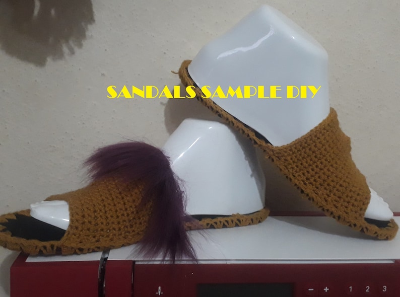 May include: A pair of brown crocheted sandals with a purple fuzzy trim. The sandals are displayed on a white mannequin. The text "SANDALS SAMPLE DIY" is visible in the image.