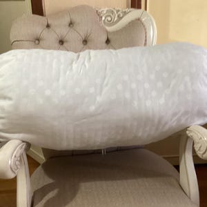 Puede incluir: Un cojín cilíndrico blanco con un sutil estampado de lunares descansa sobre una silla decorativa. La almohada es suave y de color claro y neutro. La silla tiene reposabrazos curvos y ornamentados.