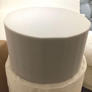 Puede incluir: Un objeto cilíndrico de espuma gris claro, posiblemente un cojín o soporte, se asienta sobre una base de tela blanca. La espuma tiene una parte superior lisa y redondeada y un color uniforme. La base de tela parece ser un material suave y texturizado.