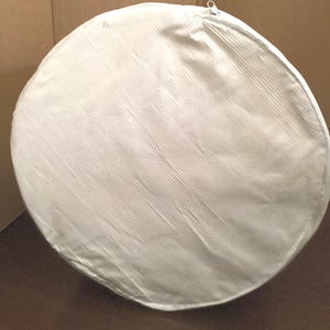 Puede incluir: Una bolsa de almacenamiento redonda blanca con cierre de cremallera. La bolsa está hecha de un tejido texturizado y tiene un forro blanco liso.