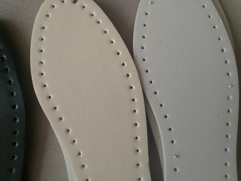 K&ouml;nnte beinhalten: Nahaufnahme von drei Schuh-Innensohlen in verschiedenen Farben: Schwarz, Creme und Grau. Jede Innensohle hat eine Reihe von gleichm&auml;&szlig;ig verteilten L&ouml;chern am Umfang, die zum N&auml;hen bestimmt sind. Die Innensohlen bestehen aus einem glatten, lederartigen Material.