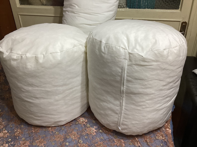May include: Three white cylindrical pillows, two of which are sitting on a patterned surface. The pillows are made of a soft, white fabric and have a zipper on the side.