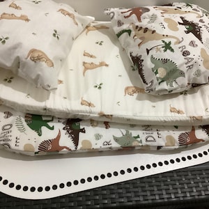 Puede incluir: Un nido de bebé blanco con temática de dinosaurios. El nido incluye una base blanca con borde perforado y varias capas de tela suave estampada. La tela presenta ilustraciones de dinosaurios, árboles y las palabras "Wild Friend" y "Roar".