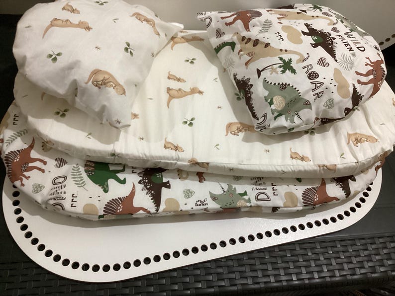 K&ouml;nnte beinhalten: Ein wei&szlig;es Babynest mit Dinosaurier-Motiv. Das Nest umfasst eine wei&szlig;e Basis mit perforiertem Rand und mehrere Schichten weichen, gemusterten Stoffes. Der Stoff zeigt Illustrationen von Dinosauriern, B&auml;umen und die Worte "Wild Friend" und "Roar".