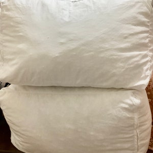 May include: Two white pillows stacked on top of each other. The pillows are made of a soft, white fabric and have a slightly wrinkled appearance.