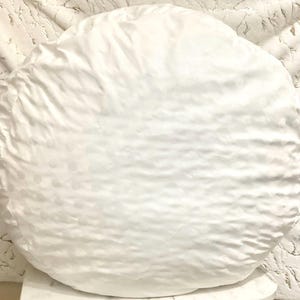 Puede incluir: Un cojín blanco grande y redondo con una superficie texturizada y un sutil estampado de lunares. El cojín está hecho de un material suave, posiblemente satinado. El color neutro sugiere que se puede utilizar como elemento decorativo o para mayor comodidad.
