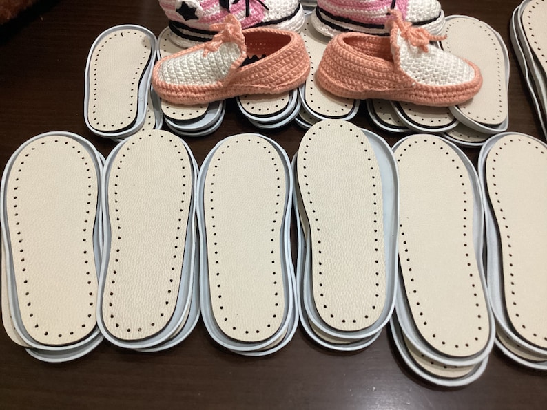 May include: A close-up of several pairs of white and pink crocheted slippers with white leather soles. The soles are stacked on top of each other.